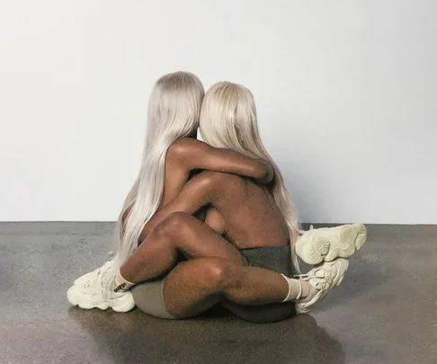 Two women with long blond hair embrace on a gray floor wearing Yeezy sneakers and minimal clothing from the Yeezy Season 6 campaign.