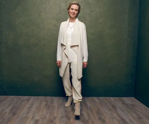 Cynthia Nixon, dressed in white attire, stands against a green textured background, posing for the camera.