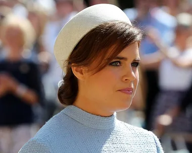 Side profile of a woman with brown hair wearing a light blue dress and a white hat, outdoors on a sunny day, blurred crowd in the background.