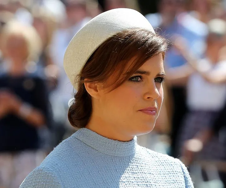 Side profile of a woman with brown hair wearing a light blue dress and a white hat, outdoors on a sunny day, blurred crowd in the background.