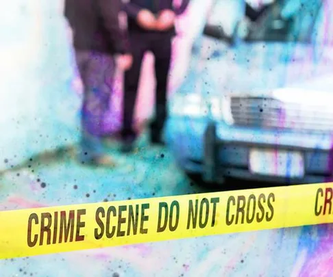 "Crime scene tape reading 'CRIME SCENE DO NOT CROSS' in front of a blurred background with two people and a car."