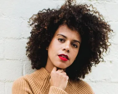 A person with curly hair and red lipstick wears a beige sweater, posing thoughtfully against a light brick wall.