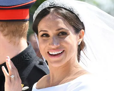 A smiling bride in a white wedding dress and tiara waving, with a groom in a military uniform visible from behind.