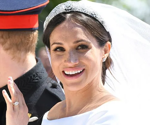 A smiling bride in a white wedding dress and tiara waving, with a groom in a military uniform visible from behind.