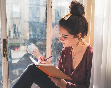 A woman with a bun writing in a notebook by a window with sunlight streaming in.