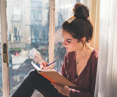 A woman with a bun writing in a notebook by a window with sunlight streaming in.