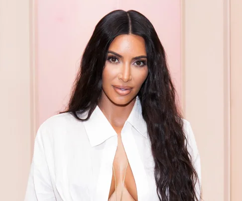 A woman with long, dark hair wearing a white, open-collared shirt, looking directly at the camera against a neutral background.