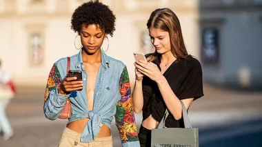 Two women standing outdoors, looking at their smartphones.