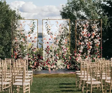 Outdoor wedding setup with rows of chairs facing a floral backdrop of flowers on glass panels, with trees and a cloudy sky in the background.