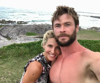 A couple taking a selfie at the beach, with rocky coastline and ocean waves in the background.