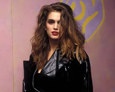 Woman with voluminous hair and red lipstick wearing a black outfit, standing against a pink and yellow abstract background.