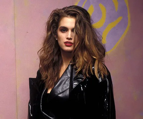 Woman with voluminous hair and red lipstick wearing a black outfit, standing against a pink and yellow abstract background.