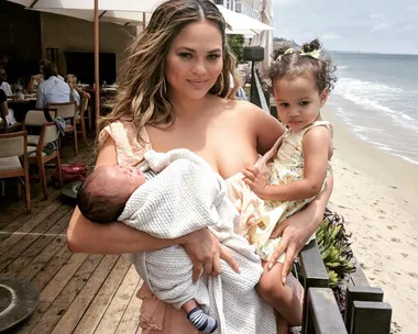 Chrissy Teigen holding her two children at a beachside restaurant, with the ocean in the background.