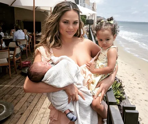 Chrissy Teigen holding her two children at a beachside restaurant, with the ocean in the background.