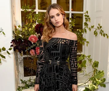 A woman in an off-shoulder black dress with geometric patterns stands by a window, surrounded by greenery and flowers.