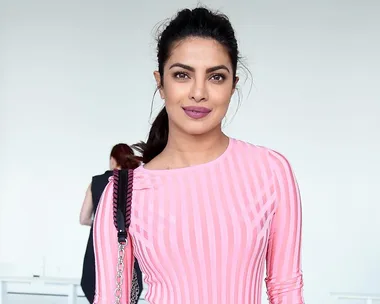 A woman wearing a pink striped top with a black shoulder bag stands against a plain background.