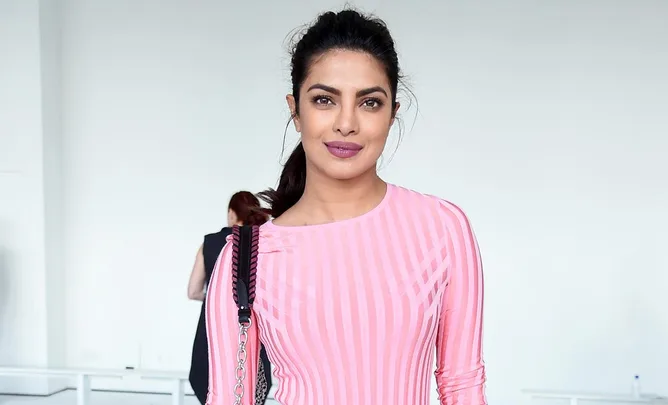 A woman wearing a pink striped top with a black shoulder bag stands against a plain background.