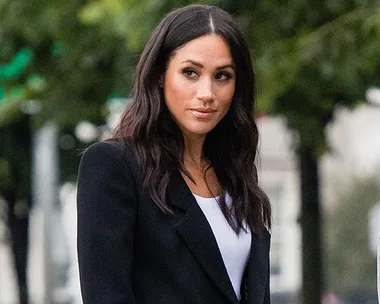 A woman with long dark hair, wearing a black blazer and white top, standing outdoors with blurred trees in the background.