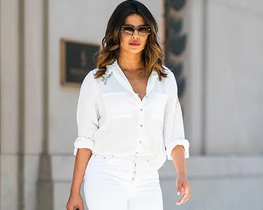 Priyanka Chopra wearing sunglasses, walks in an all-white outfit against a cityscape backdrop.