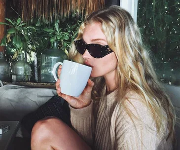 Woman with sunglasses drinking coffee from white mug in a cozy, plant-filled space.