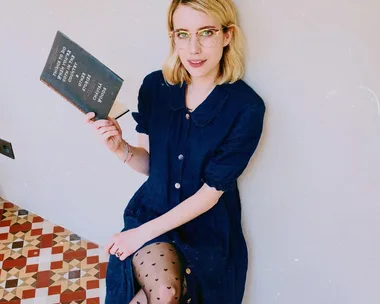 Woman in blue dress holding a book with text, sitting on a patterned floor.