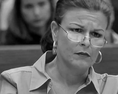 A black and white photo of a woman with glasses, looking serious in a courtroom.