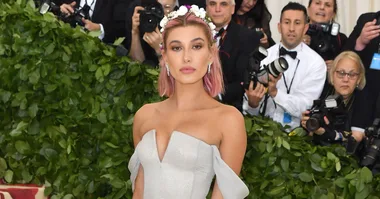 A woman in a strapless silver gown and floral headpiece standing in front of cameras and green foliage.