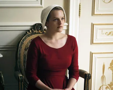 A woman in a red dress and white bonnet sits on a vintage chair, characterized by the setting from "The Handmaid's Tale."