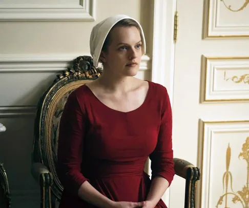 A woman in a red dress and white bonnet sits on a vintage chair, characterized by the setting from "The Handmaid's Tale."