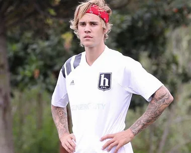 A man with a red bandana and tattooed arms wearing a white H C.F.C. shirt, standing outdoors with his hands on his hips.