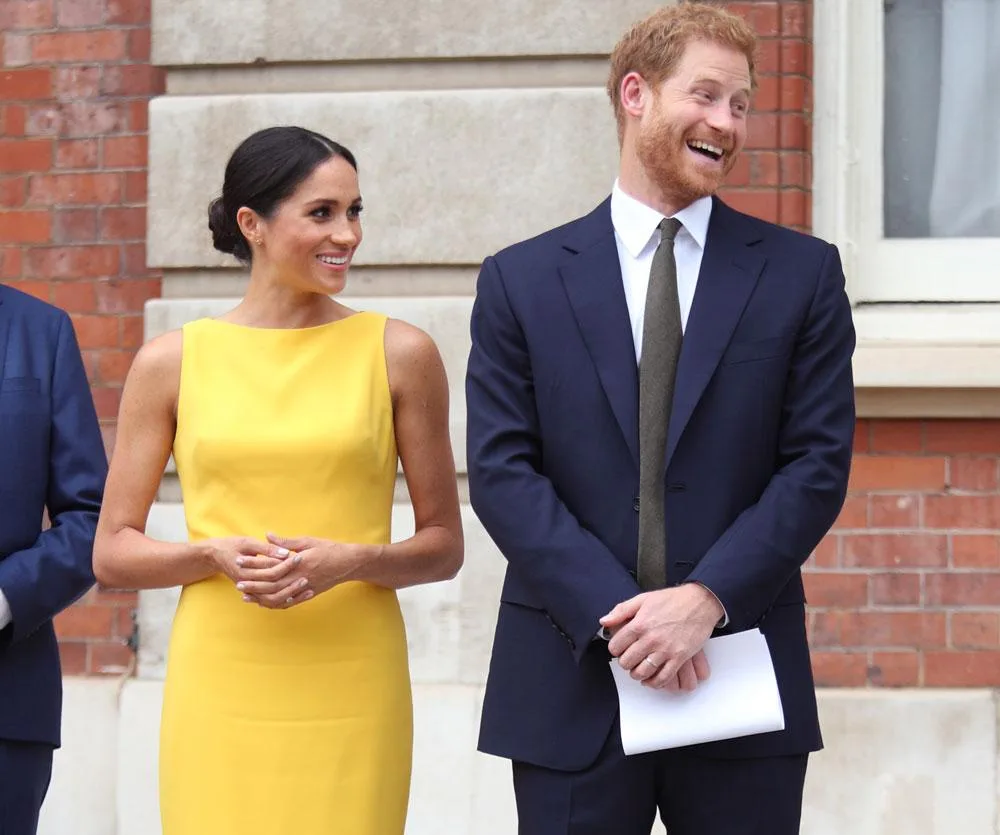 Prince Harry and Meghan Markle smiling and standing together outside a building. She wears a yellow dress; he is in a navy suit.