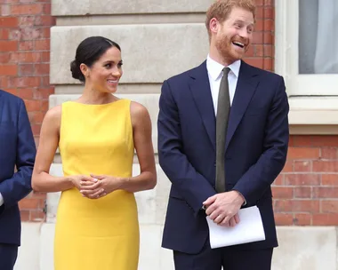 Prince Harry and Meghan Markle smiling and standing together outside a building. She wears a yellow dress; he is in a navy suit.