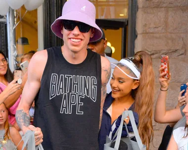 Pete Davidson in a purple bucket hat and tank top beside Ariana Grande with a visor and ponytail, smiling and holding bags.