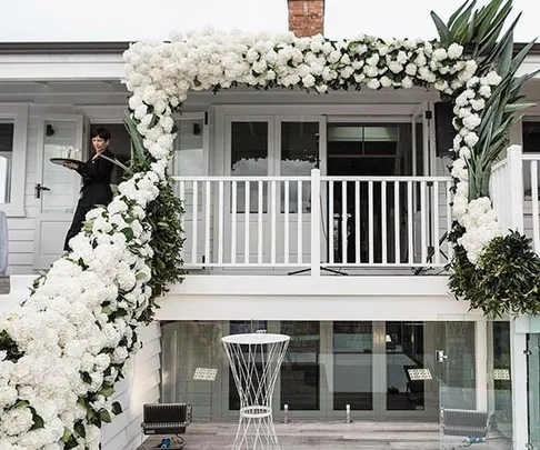 White floral wedding arch decorates a house balcony with a waiter on the left carrying a tray.