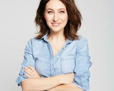 Woman in blue denim shirt smiling with arms crossed against a plain background.