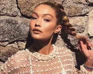 A person with braided hair poses against a stone wall wearing a lacy top, looking confidently at the camera.