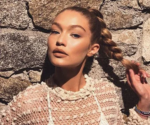 A person with braided hair poses against a stone wall wearing a lacy top, looking confidently at the camera.