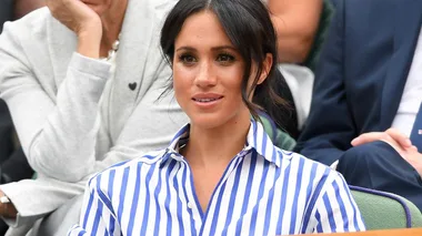 Meghan Markle seated at Wimbledon, wearing a blue and white striped shirt, with spectators in the background.