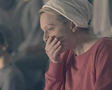 June Osborne in "The Handmaid's Tale" dressed in a red outfit, covering her mouth with her hand, looking concerned.