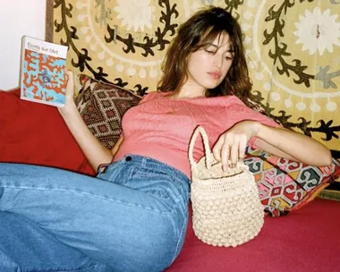 Woman lounging with a book titled "Écrits sur l'Art" and holding a woven handbag, against a patterned tapestry backdrop.