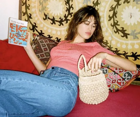 Woman lounging with a book titled "Écrits sur l'Art" and holding a woven handbag, against a patterned tapestry backdrop.