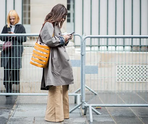 Woman with brown coat and orange bag stands by metal barricade using her phone; another woman in grey coat in background, also on phone.