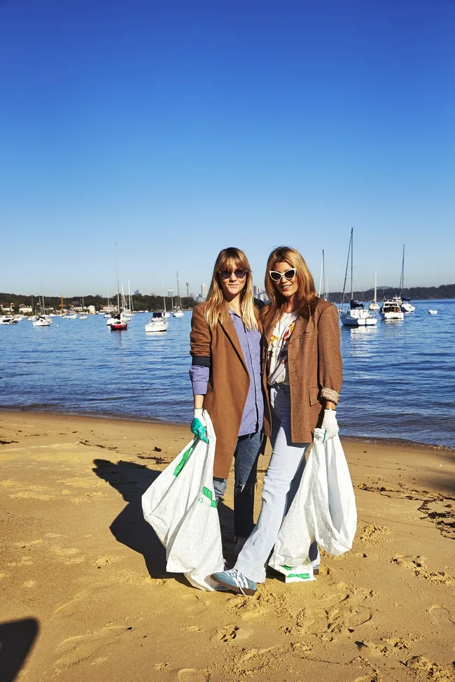 Two women wearing sunglasses and brown coats cleaning up a beach with bags, boats in the background.