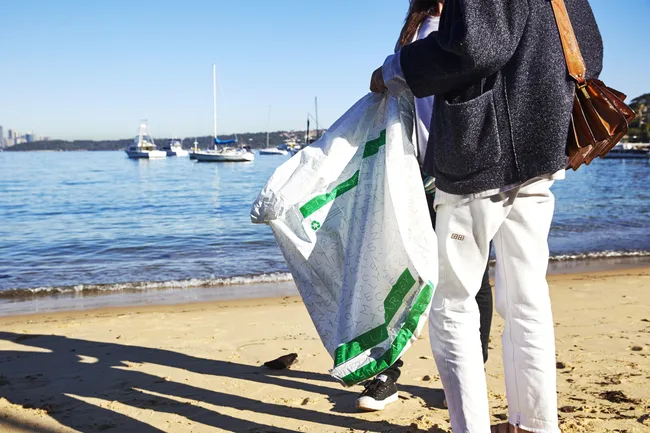 Two people on a beach holding a large white bag with green trim; boats are seen on the water in the background.