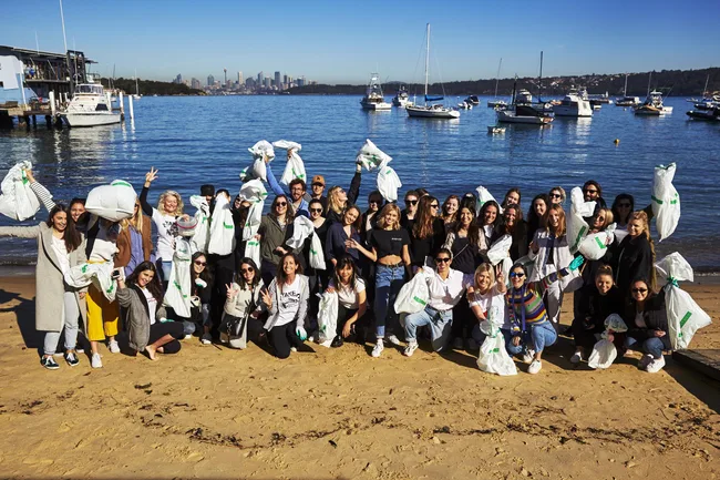 Group of people on a beach holding white bags with boats and city skyline in the background.