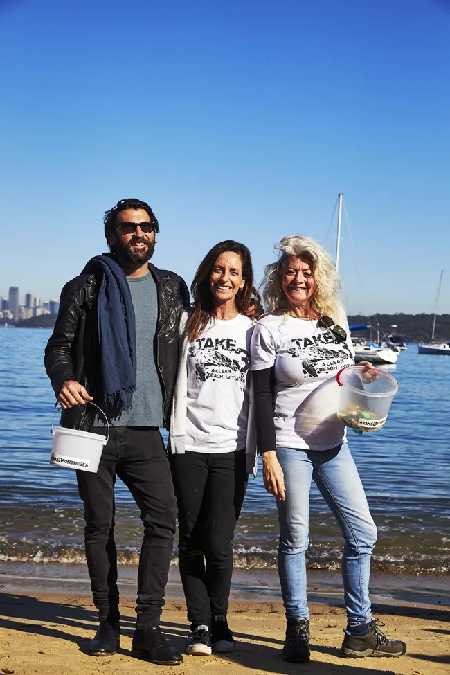 Three people smiling on a sandy beach, holding buckets, with a blue sky and boats in the background.