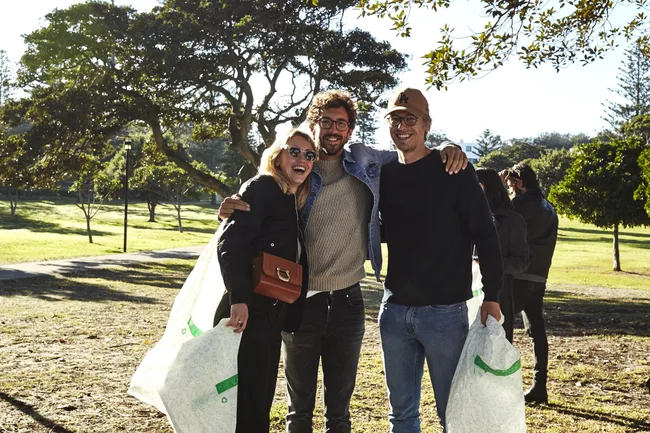 Group of three people smiling outdoors, holding recycling bags in a park setting with trees and grass.