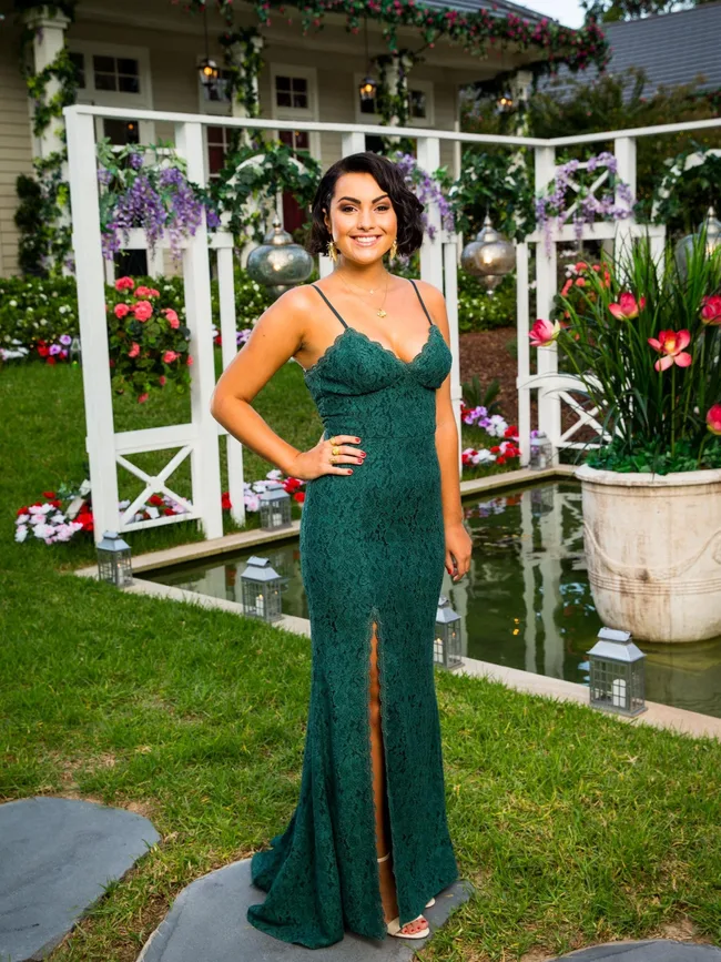 A woman in a green lace dress stands smiling in a garden with flowers and lanterns, in front of a decorated house.