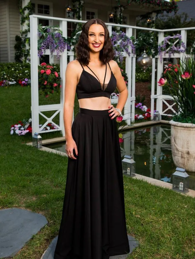 Woman in a black two-piece evening gown stands in a garden setting with flowers and white trellis backdrop.