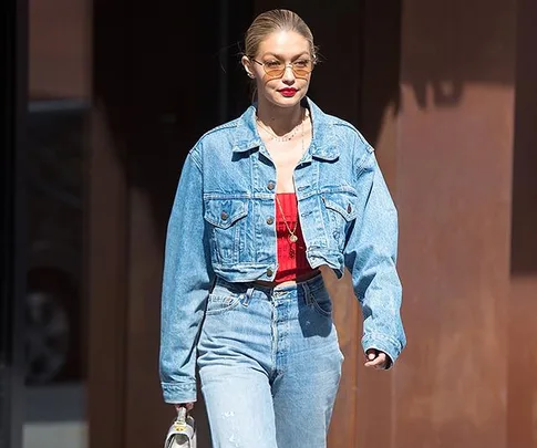 A person in a cropped denim jacket and jeans walks outside wearing sunglasses and holding a small bag.